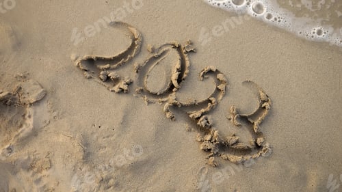 Preview: Closeup of female finger writing 2023 year on sea beach next to rolling waves