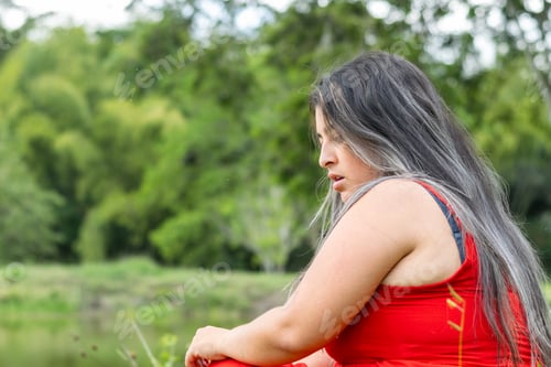 Preview: woman looking down at the ground with a worried face, sitting with a forest in the background.