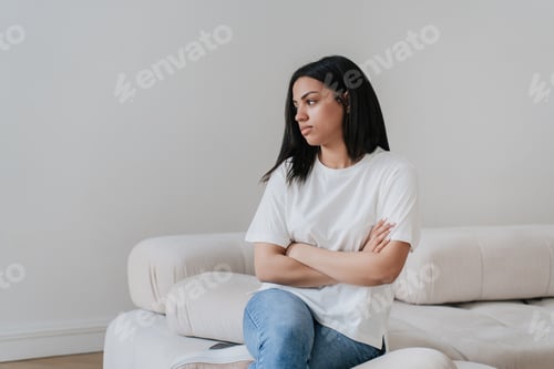 Preview: Upset afro Brazilian young woman sitting on sofa home alone. African american girl in white t- shirt