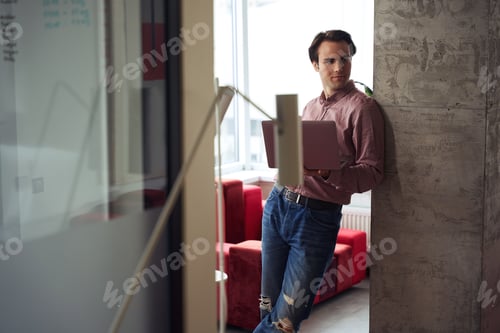Preview: Man Leans Against Wall Holding Laptop in Workplace
