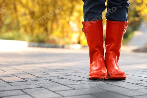 Preview: Woman wearing red rubber boots on paved street in park, closeup. Autumn walk