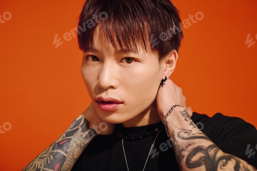 Preview: Stylish young asian man poses against a vibrant orange backdrop while showcasing unique tattoos