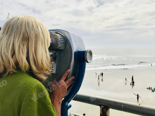 Preview: A blonde baby boomer woman looks through a telescope on the beach