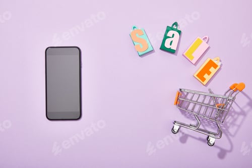 Preview: top view of smartphone near shopping bags with sale lettering falling into toy cart on violet