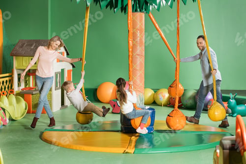 Preview: smiling parents looking at happy children swinging on swings in entertainment center