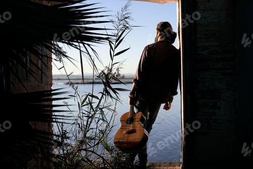 Preview: caucasian boy with his back relaxed by the lake leaning on the door of an old building with a guitar