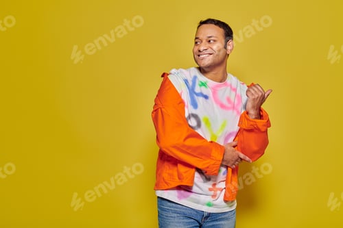 Preview: positive indian man in orange jacket and diy t-shirt pointing with thumb on yellow background