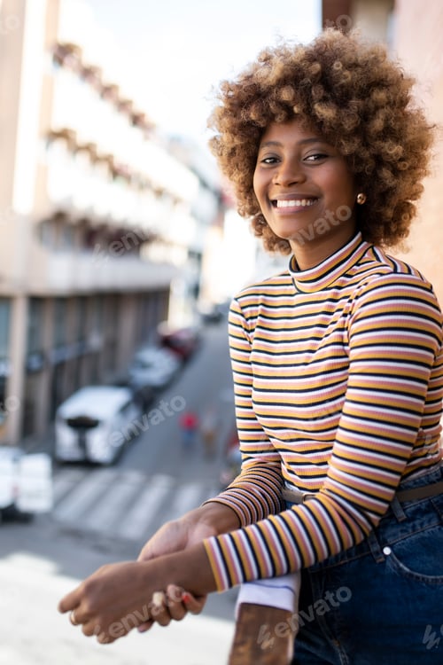 Preview: Portrait of smiling woman standing in a balcony with city background. Girl staring at camera with a