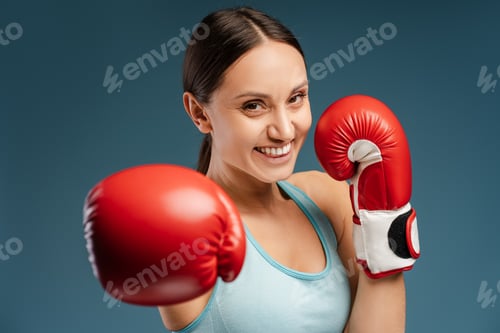 Preview: Beautiful smiling woman wearing red boxing gloves, doing punch isolated on blue background