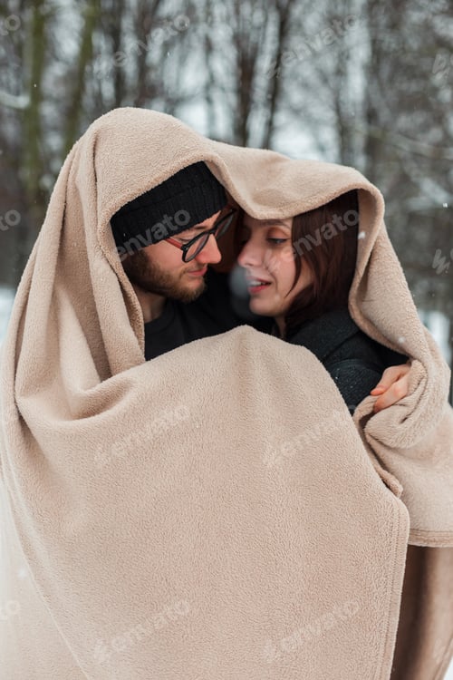 Preview: Happy beautiful couple of lovers wrapped in a beige blanket in the park on a winter day with snow