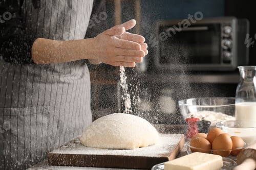 Preview: Woman sprinkling flour over dough on table in kitchen