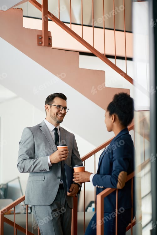 Preview: Happy businessman talking to black female colleague while drinking coffee in a hallway.