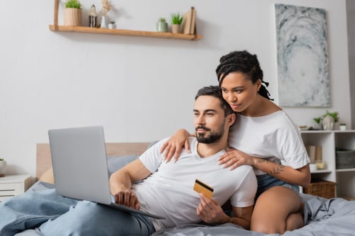 young african american woman hugging shoulders of bearded man with laptop and credit card on bed