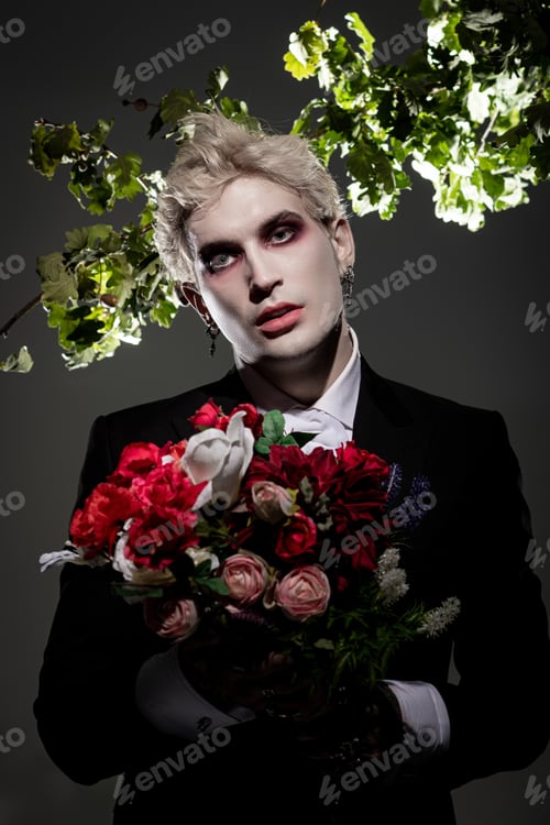Preview: Young man in stunning Halloween costume holds a bouquet against a dark backdrop