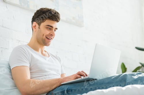 Preview: handsome man in white t-shirt and jeans sitting on bed, smiling and typing on laptop in bedroom