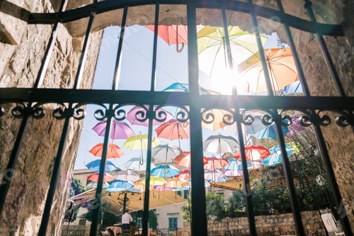 Preview: View through a wrought-iron fence to colorful umbrellas hanging above the street of an ancient town