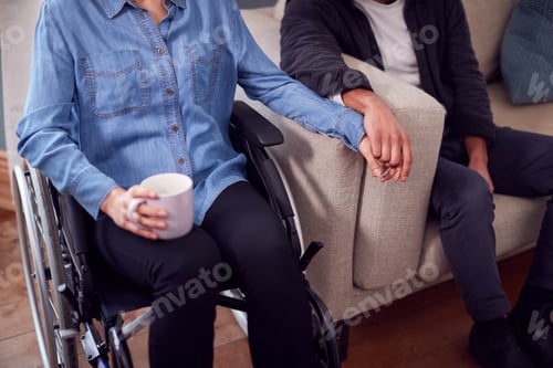 Preview: Close Up Of Couple At Home With Disabled Woman In Wheelchair Holding Hands With Man Sitting On Sofa