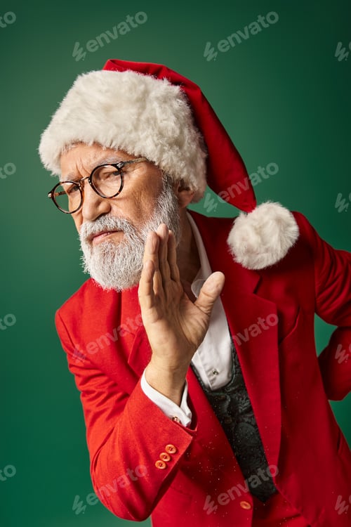 Preview: serious white bearded Santa gesturing on dark green backdrop looking at camera, winter concept