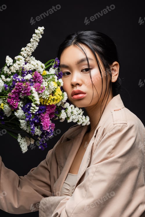 Preview: Young asian model in trench coat holding wildflowers near face isolated on black