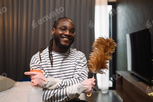 Preview: Happy smiling young African guy holding a feather duster ready for cleaning his apartment