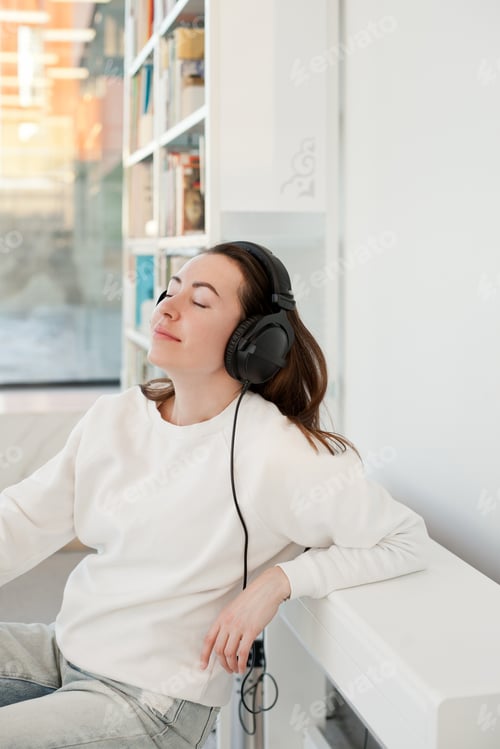 Preview: Young woman in white sweatshirt listening to music with headphones at a public library eyes closed