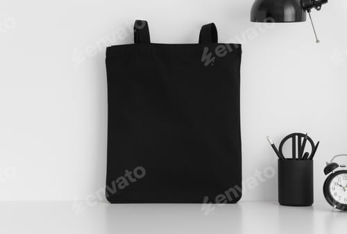 Preview: Black tote bag mockup with workspace accessories on a white table.