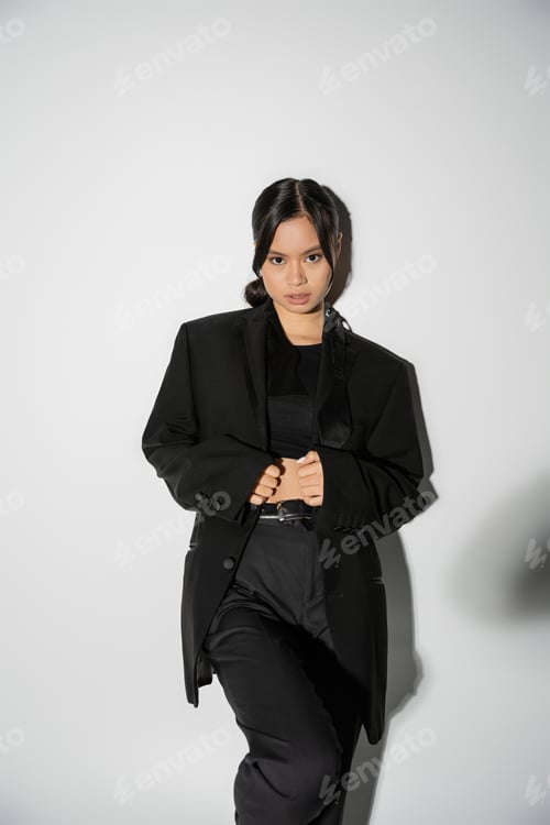 Preview: brunette asian woman in black elegant suit looking at camera on grey background