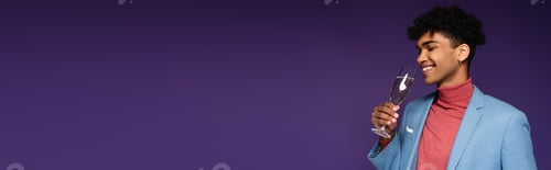 Preview: curly african american man in blue blazer holding glass of champagne while smiling on purple, banner