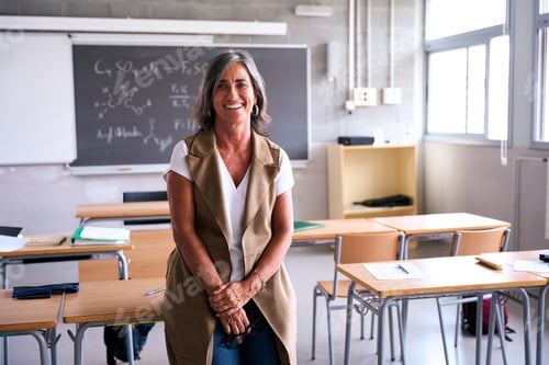 Preview: Middle-aged female high school teacher looking smiling at camera with joined hands in classroom.