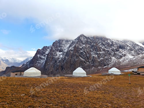 Preview: Yurts on a mountain plateau with snowy peaks in the background, under a cloudy sky