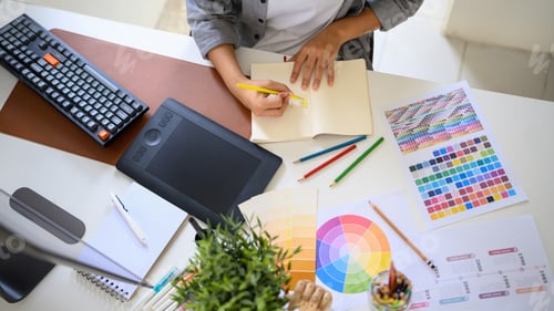 Preview: Top view creative woman sketching logo ideas in notebook with color samples and pencils