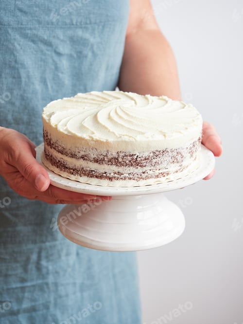 Preview: crop woman with cake stand in her hands with layered chocolate cake decorated with white buttercream