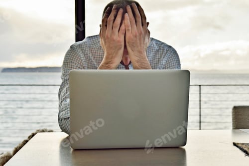 Preview: Man sitting with hands over face with laptop computer on table.