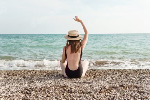 Preview: A woman in a black bathing suit and hat on the beach, on a rocky beach.