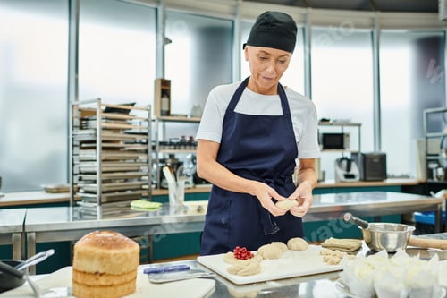 Preview: attractive mature woman in blue apron and toque working with dough on kitchen, confectionery