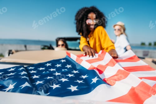 Preview: selective focus of multiethnic young women resting in car with american flag at seaside