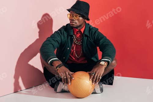 Preview: fashionable young african american man sitting while holding golden basketball ball and looking away