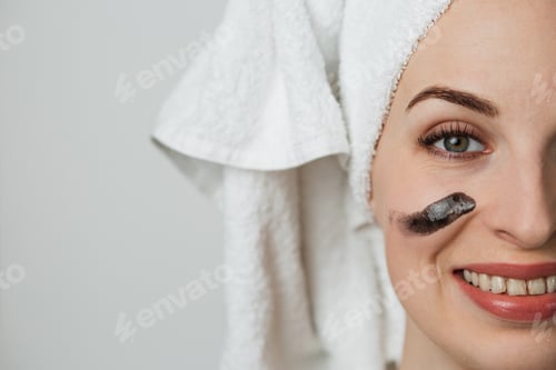 Preview: Half face portrait of caucasian young lady applying black face cream over grey background.