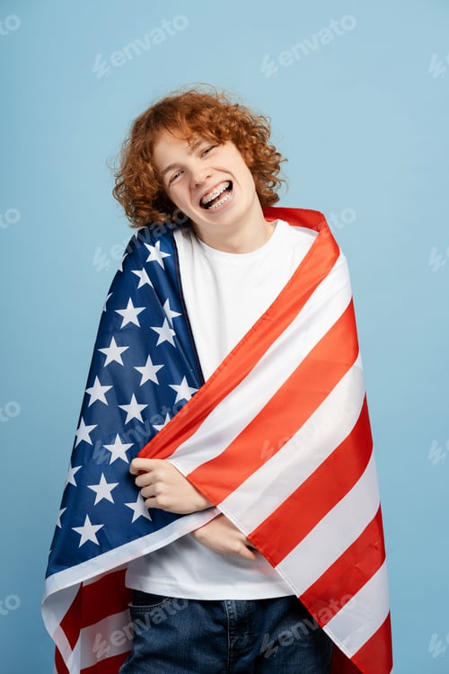 Preview: Happy red haired teen boy with braces, wrapped in large American flag, isolated on a blue background