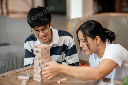 Preview: Two cheerful Asian friends enjoy playing Jenga, focusing on removing a wooden block.