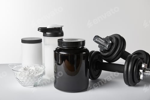 Preview: heavy dumbbells near jars and sports bottle with protein shake on white