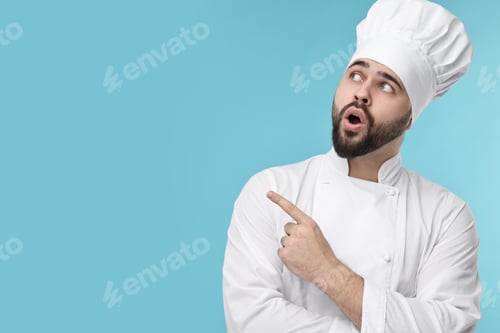 Preview: Surprised young chef in uniform pointing at something on light blue background. Space for text