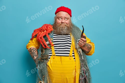 Preview: Pensive sailor with thick beard enjoys smoking tobacco pipe and focused somewhere dressed in overall
