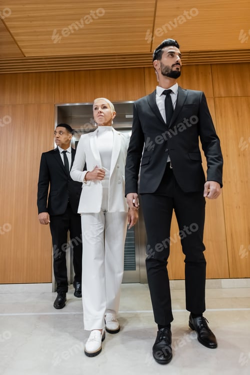 Preview: full length view of stylish senior business lady walking near private interracial bodyguards