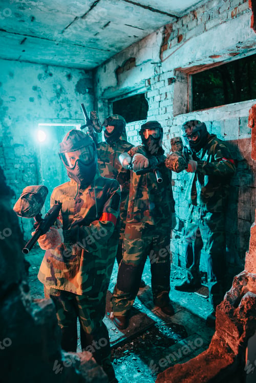 Preview: paintball team in uniform and protective masks aiming by paintball guns at camera in abandoned