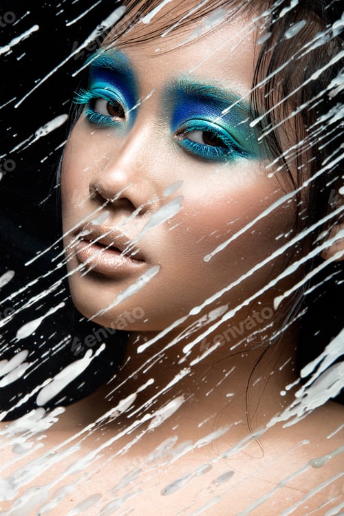 Preview: Beautiful Asian girl with bright blue make-up behind glass and drops of wax. Beauty face.
