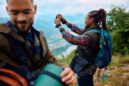 Preview: Happy black woman photographing nature with cell phone while hiking with her friend.