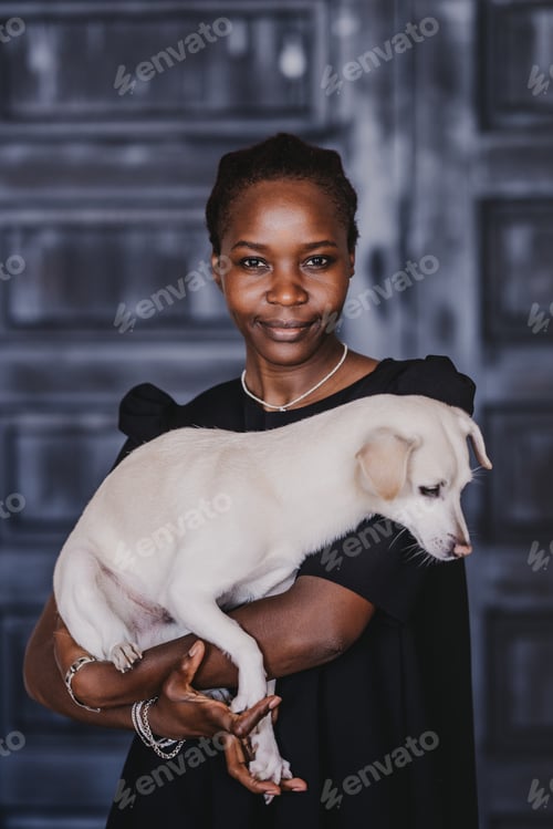 Preview: Vertical shoot of positive African young woman in black dress carrying dog on hands looks at camera.
