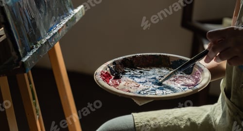 Preview: Close-up photo of female artist mixing acrylic paints on a palette. Canvas in the drawing room