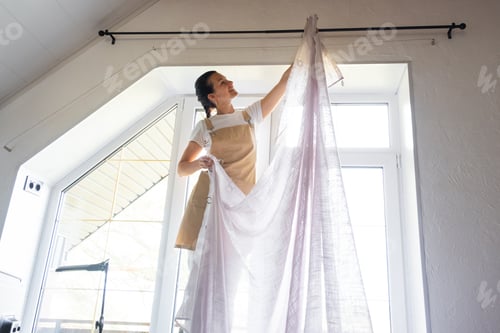 Preview: Woman in apron hangs transparent tulle curtains on large attic windows in the house inside